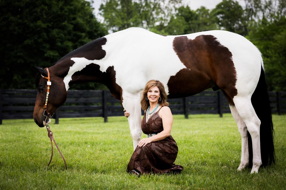 A beautiful black and white paint horse grazes next to a woman in a brown dress.
