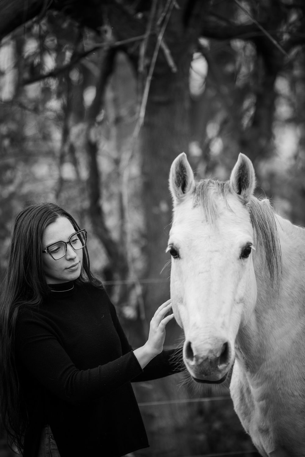 A gentle moment between a white horse and someone in black against a moody forest backdrop.