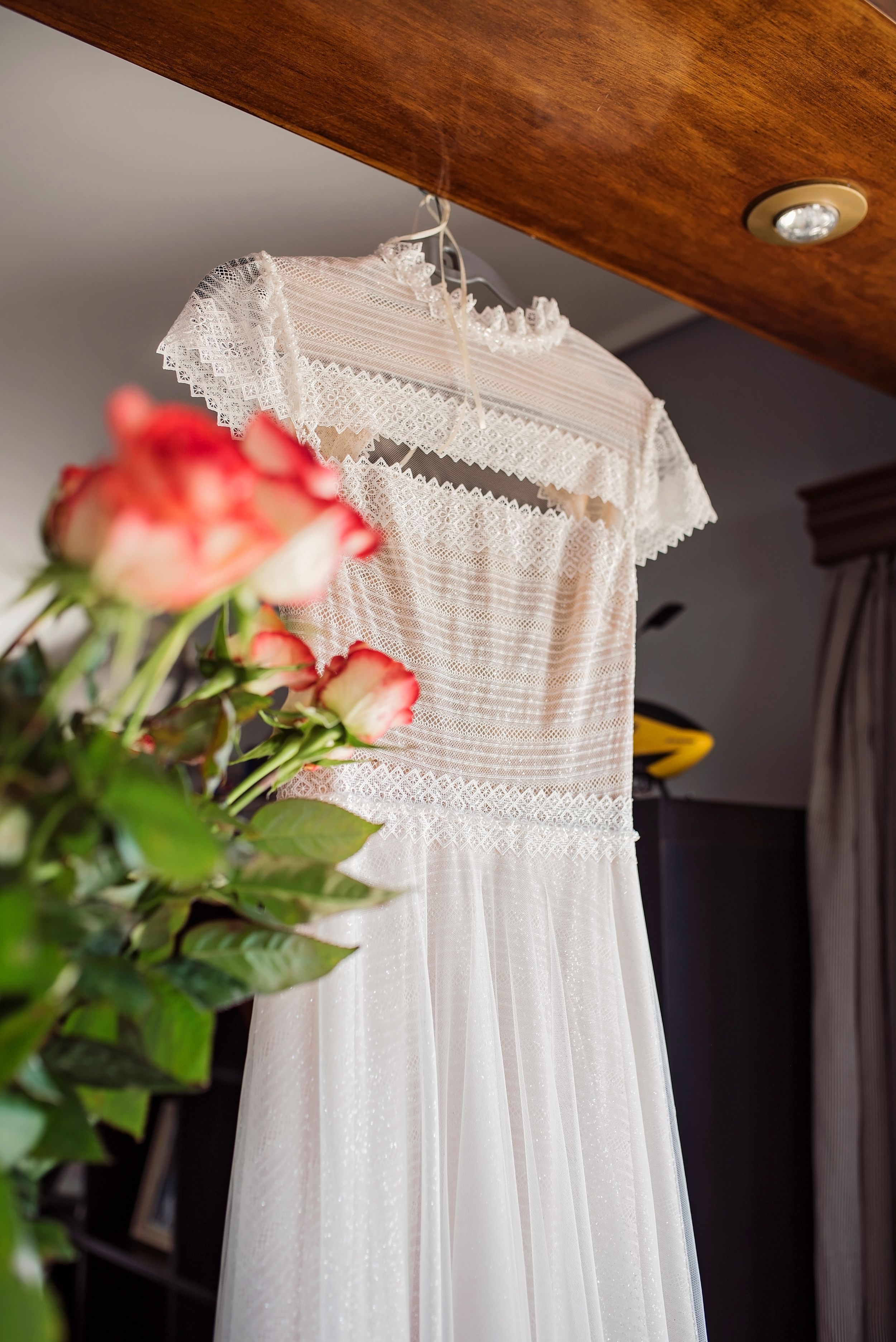 A delicate white lace wedding dress hangs against a wooden ceiling with coral roses in the foreground.