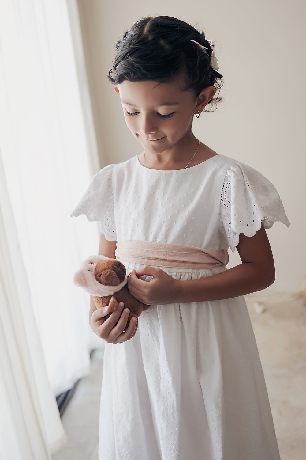 A child wearing a white eyelet lace dress holds a small teddy bear while standing by a window.