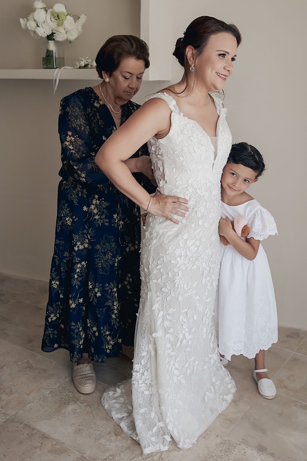 A wedding dress fitting scene showing a lace gown being adjusted with a navy floral dress and white sundress nearby. Alquimia Tulum