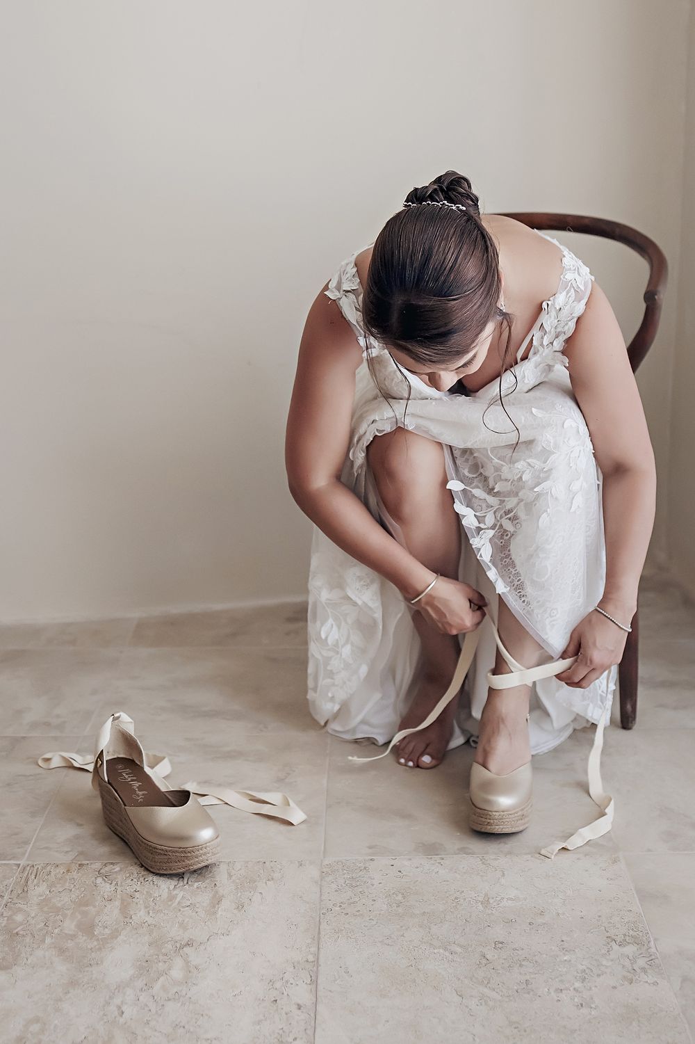 Someone in a white lace wedding dress fastening shoes while sitting on a wooden chair.
