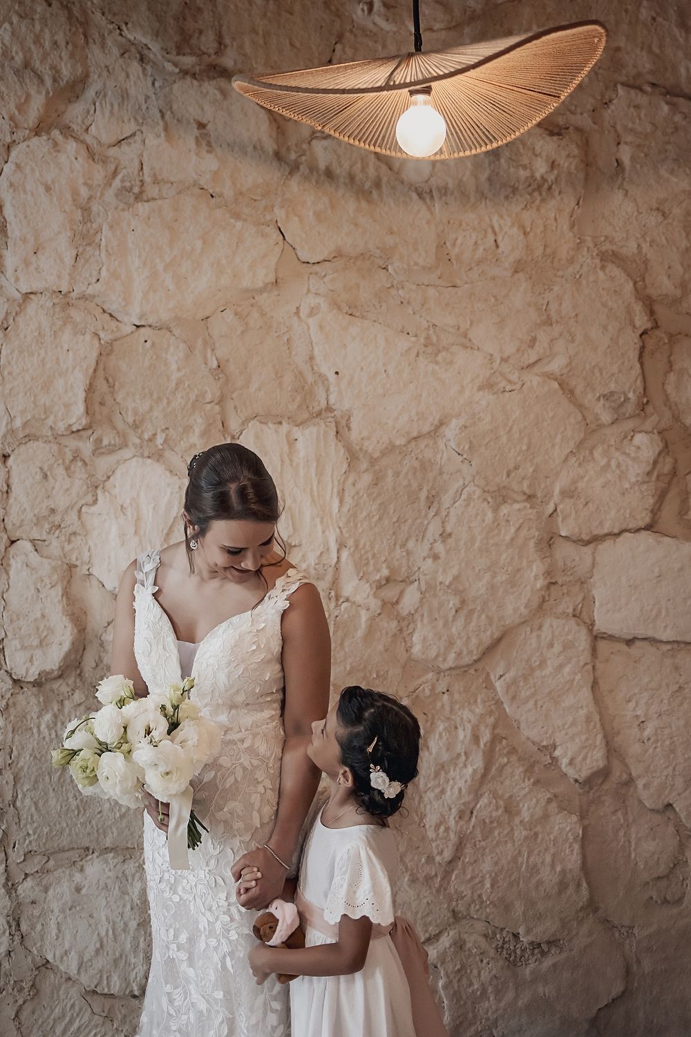 A tender moment captured between two figures in white dresses against a rustic stone wall beneath a stylish pendant light. Alquimia Tulum
