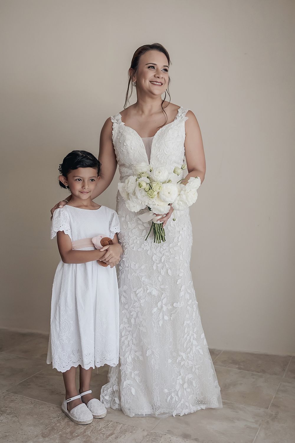 A bride in a white lace gown stands with a young attendant in a white dress, both smiling and holding bouquets.