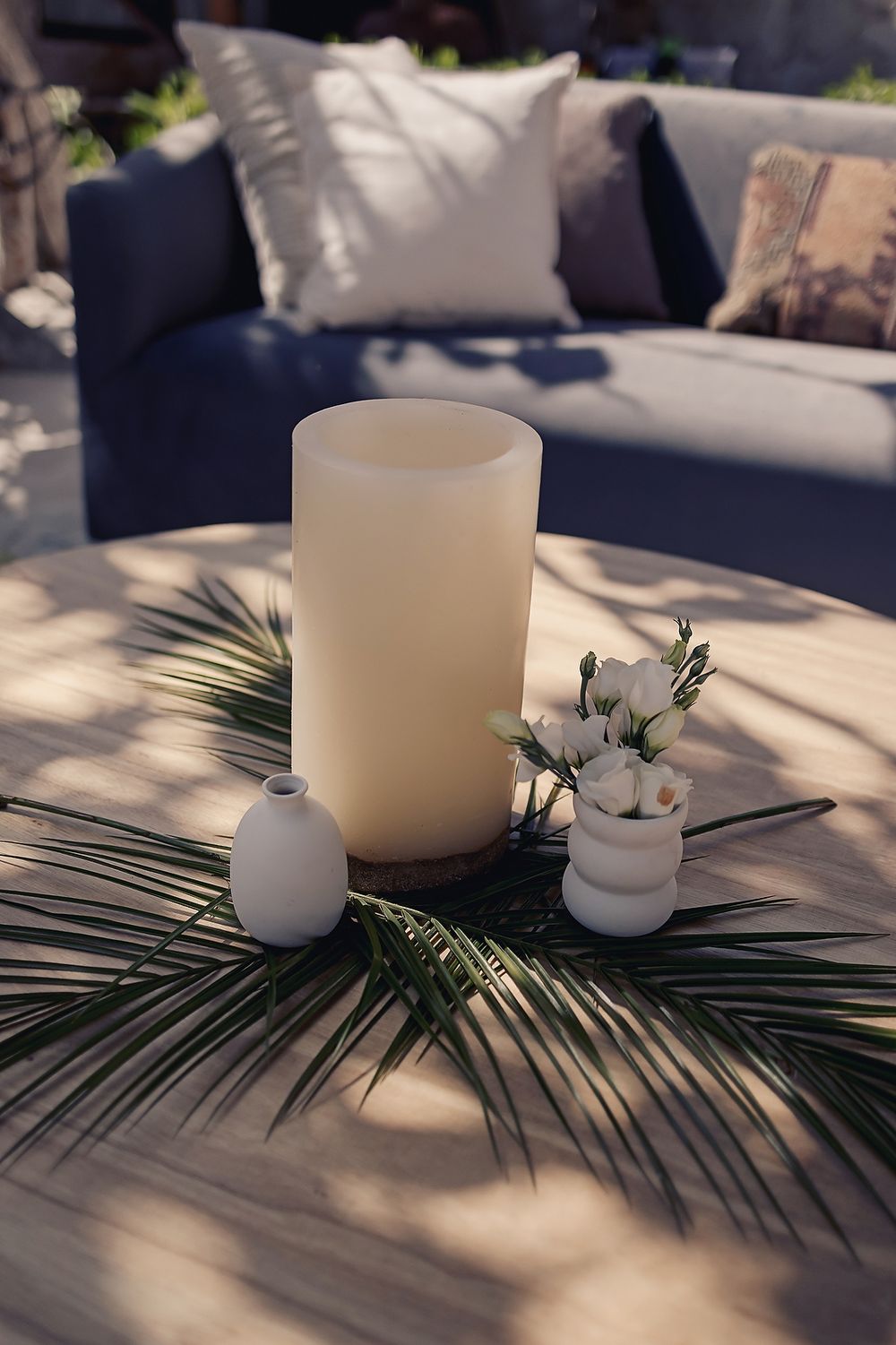 Elegant white pillar candle and small ceramic vases arranged with palm fronds on a light surface near a navy couch.