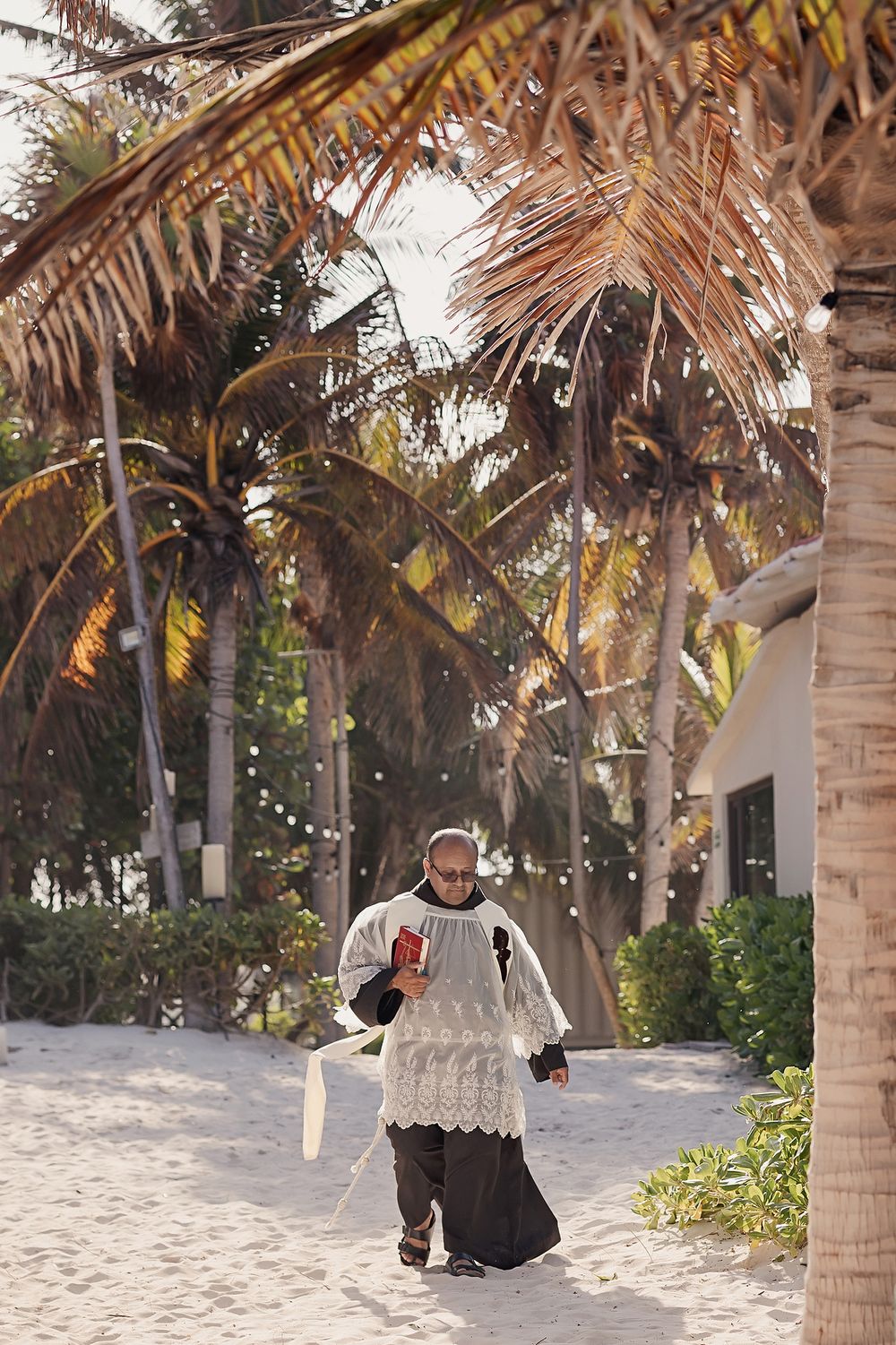 A person wearing a white ceremonial robe walks under tropical palm trees on a sandy path.