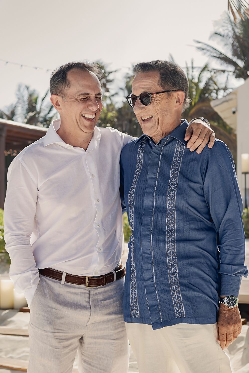 Two men in casual attire share a heartfelt moment outdoors with palm trees in the background.