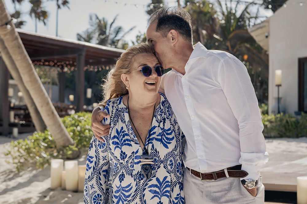 A couple shares a joyful moment outdoors, with one wearing a patterned blue dress and sunglasses.