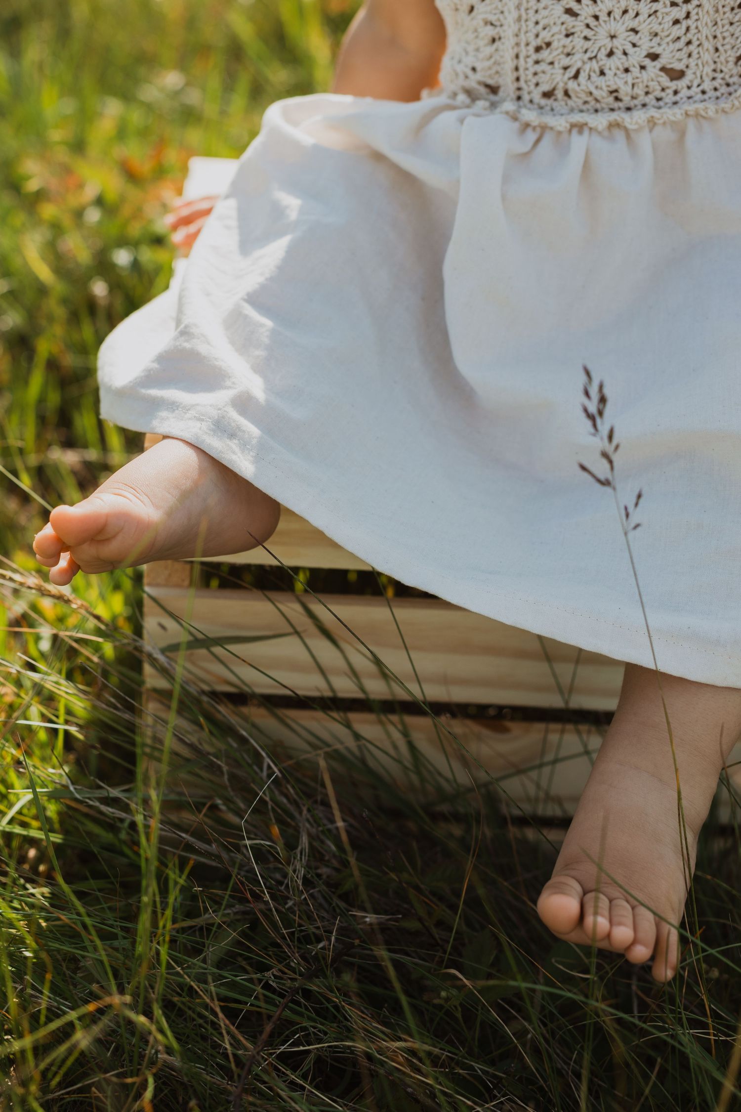 Bare feet in a flowing white dress rest delicately on a wooden surface amidst green grass and wildflowers.