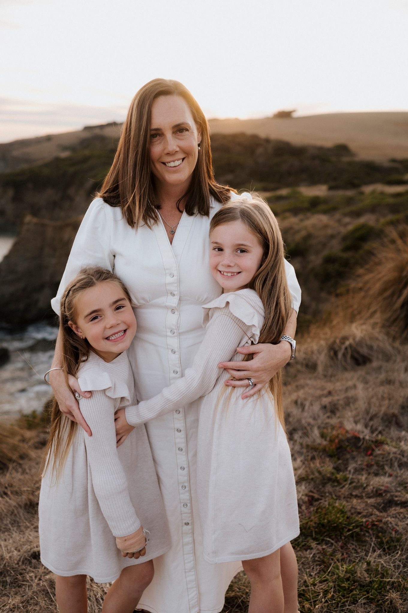 Family in white dresses poses for portrait on coastal cliffs at golden hour.