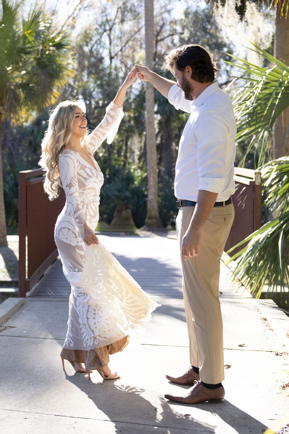 A couple in light-colored wedding attire dancing together on a sunlit boardwalk surrounded by palm trees and forest.