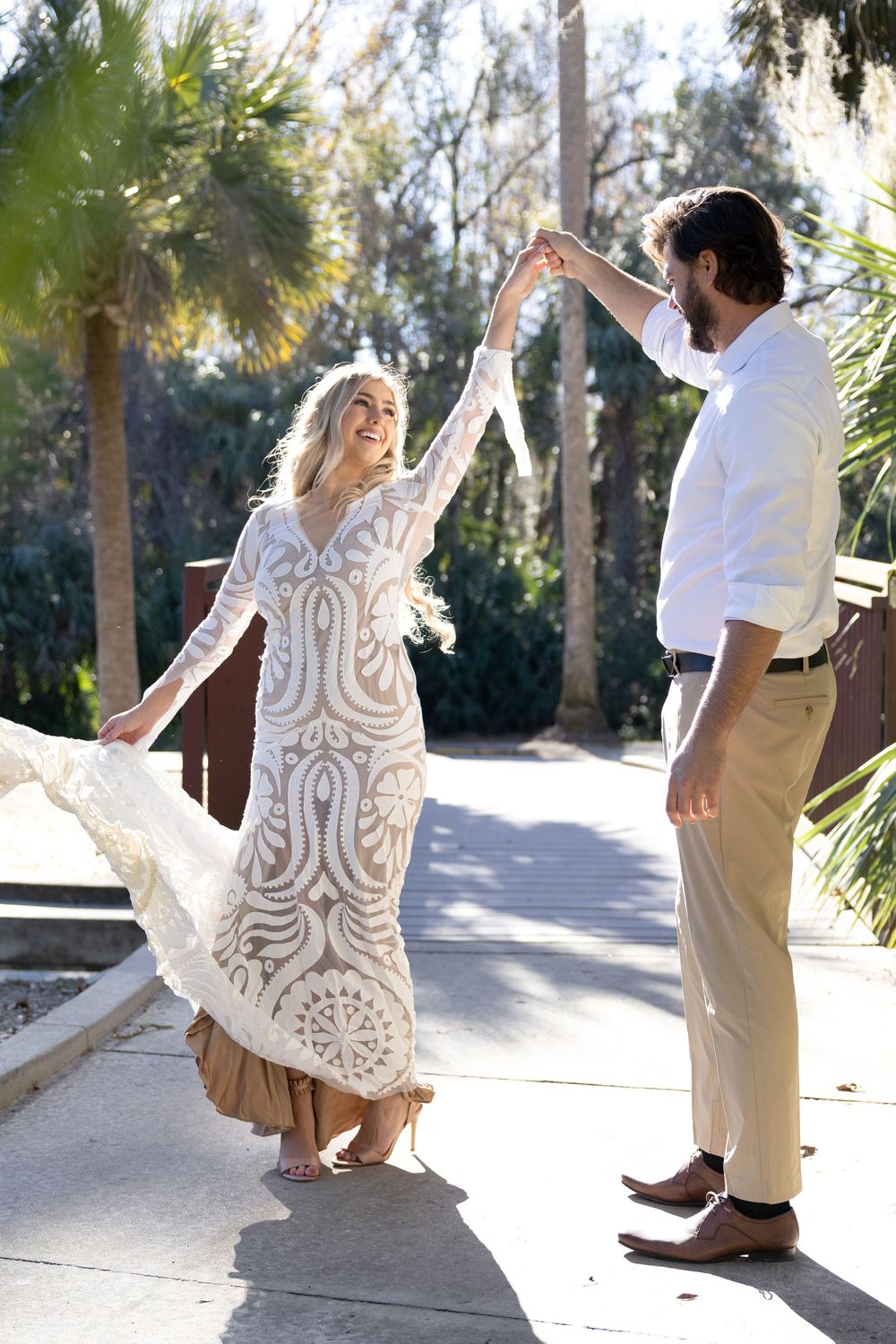 A couple dancing outdoors with palm trees, the woman wearing a flowing white lace dress and the man in a white shirt.