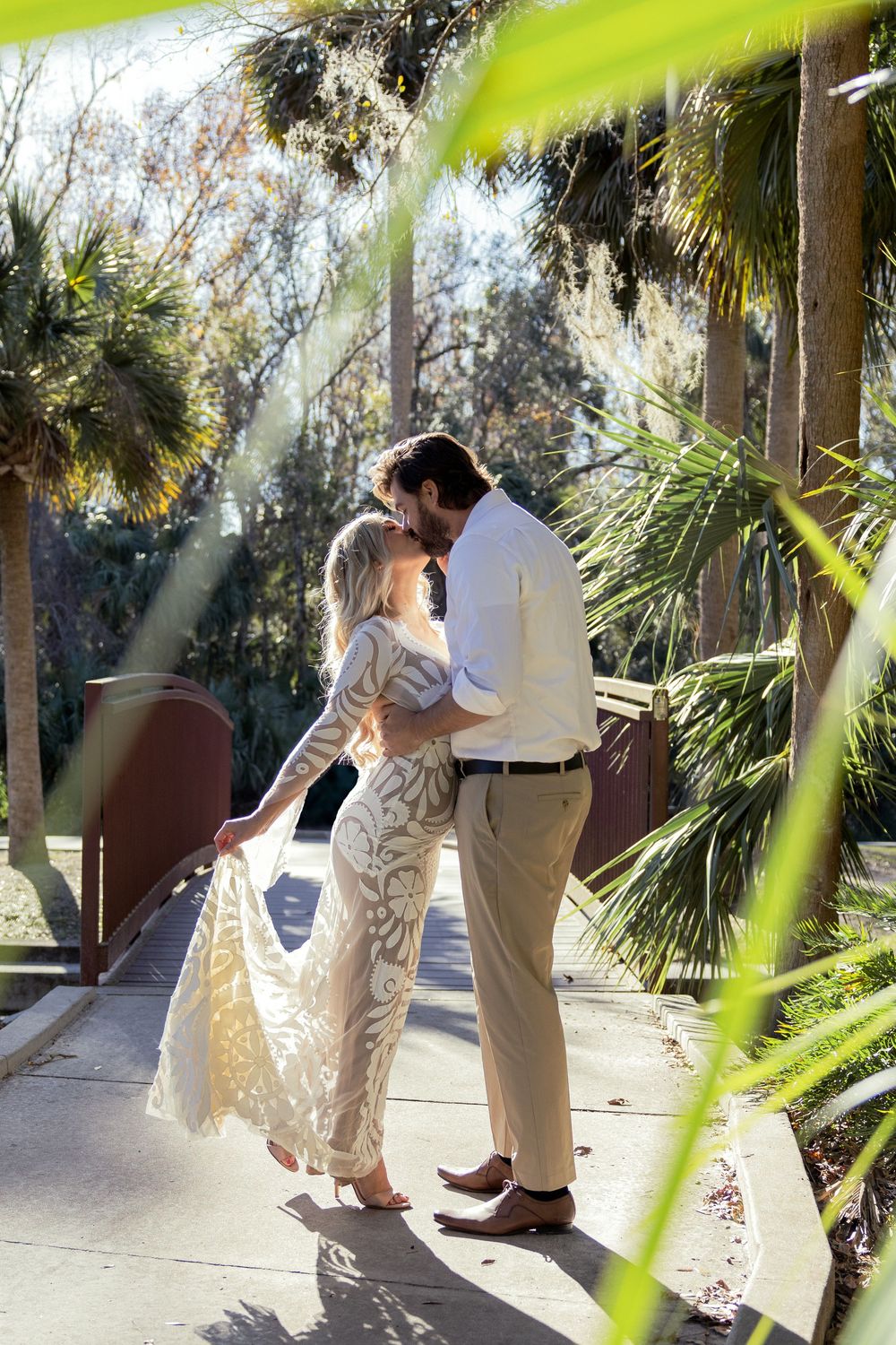 A romantic couple shares an intimate moment on a walkway surrounded by tropical palm trees in dappled sunlight.