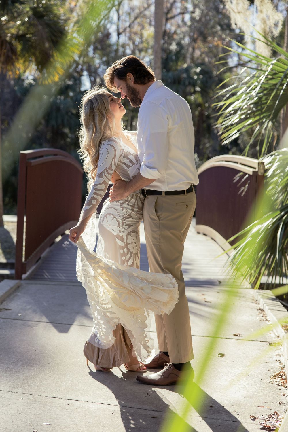 A romantic wedding couple embraces on a wooden boardwalk surrounded by tropical palm fronds in golden sunlight.