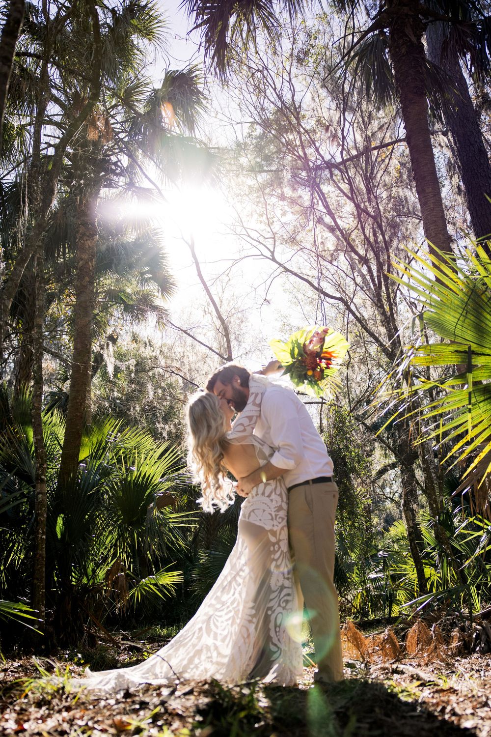 Couple sharing romantic kiss in sunlit forest with palm trees and streaming sunbeams creating magical atmosphere.