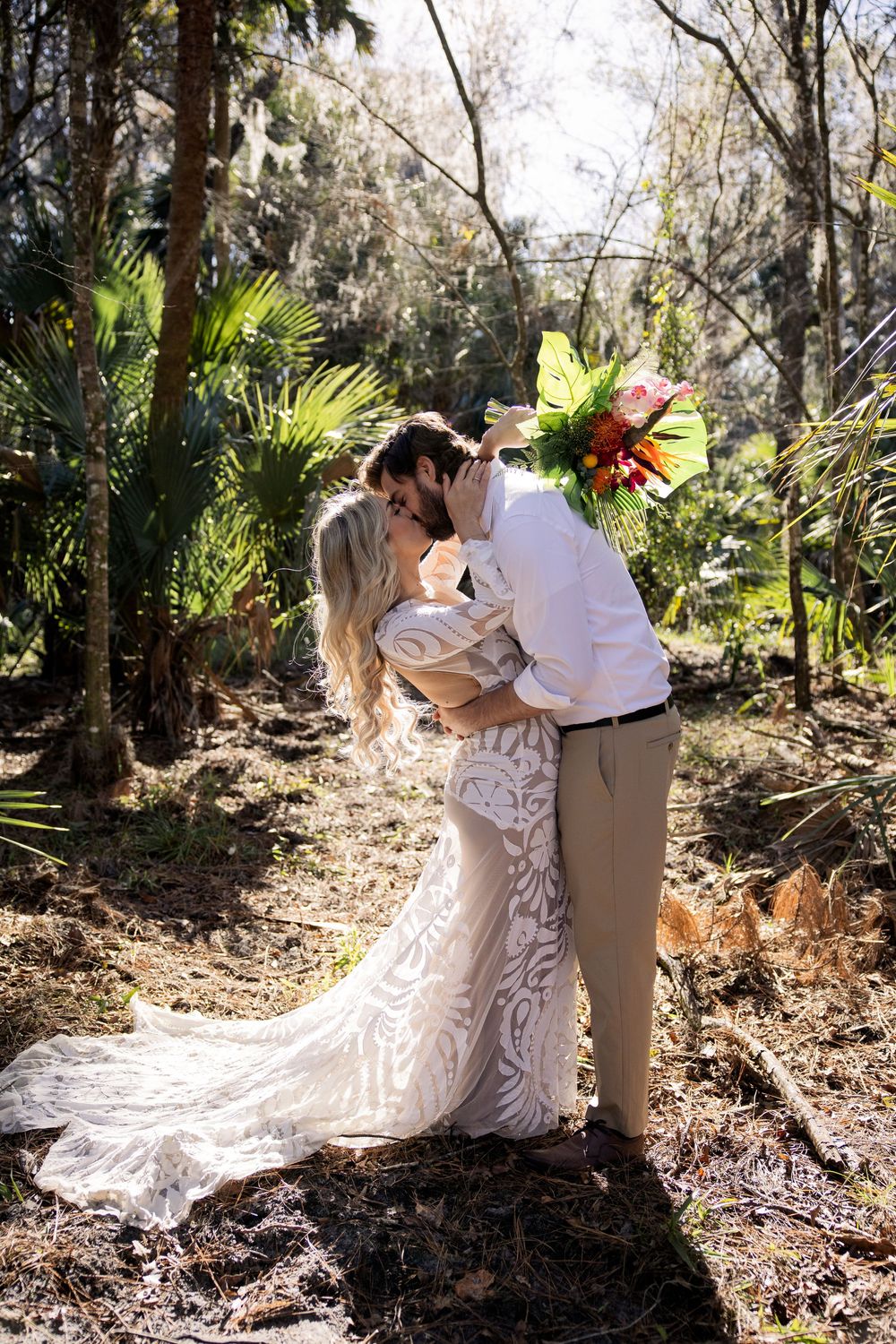 Two people share a romantic kiss among palm trees and sunlit foliage in a lush garden setting.