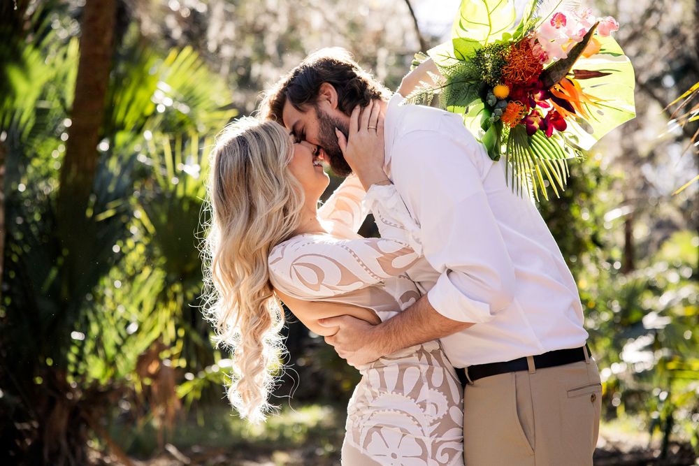 A couple in elegant white attire share an intimate kiss in a lush garden setting with bright floral decorations.