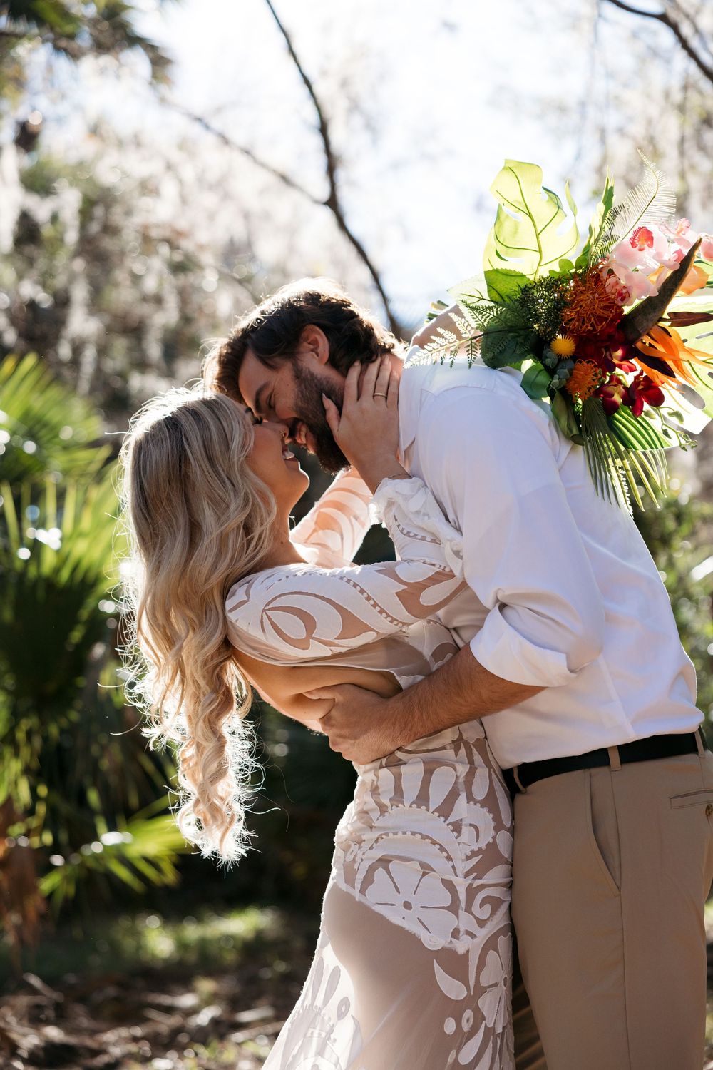 A romantic wedding couple embraces in a sun-drenched garden setting with tropical foliage and vibrant florals.