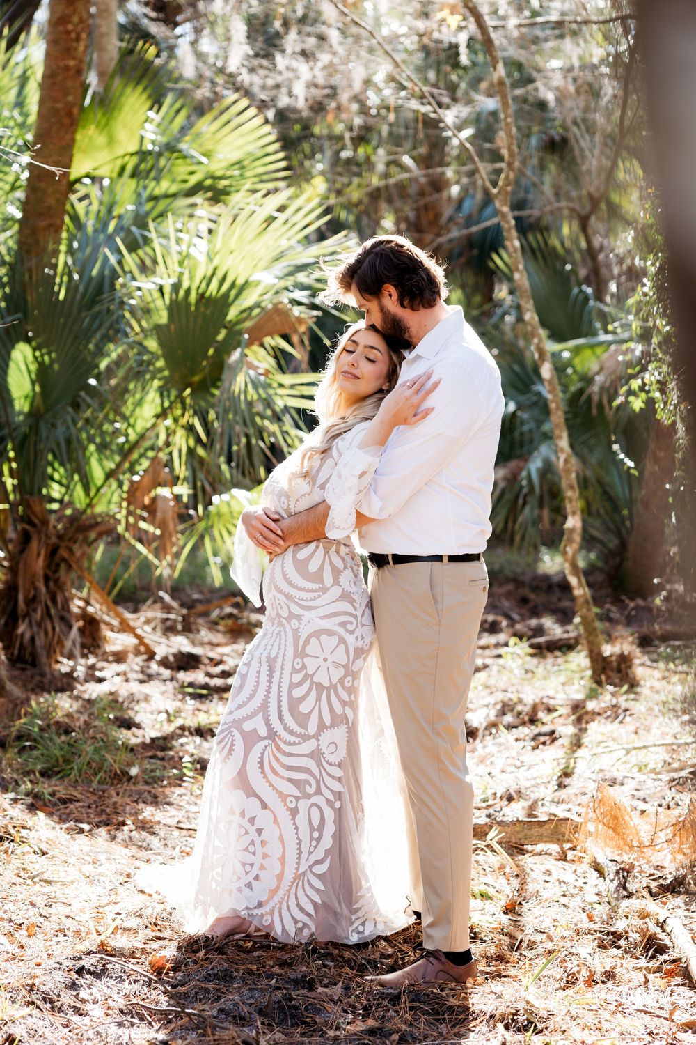 Couple embraces in a tropical garden setting with palm trees and dappled sunlight creating a romantic atmosphere.