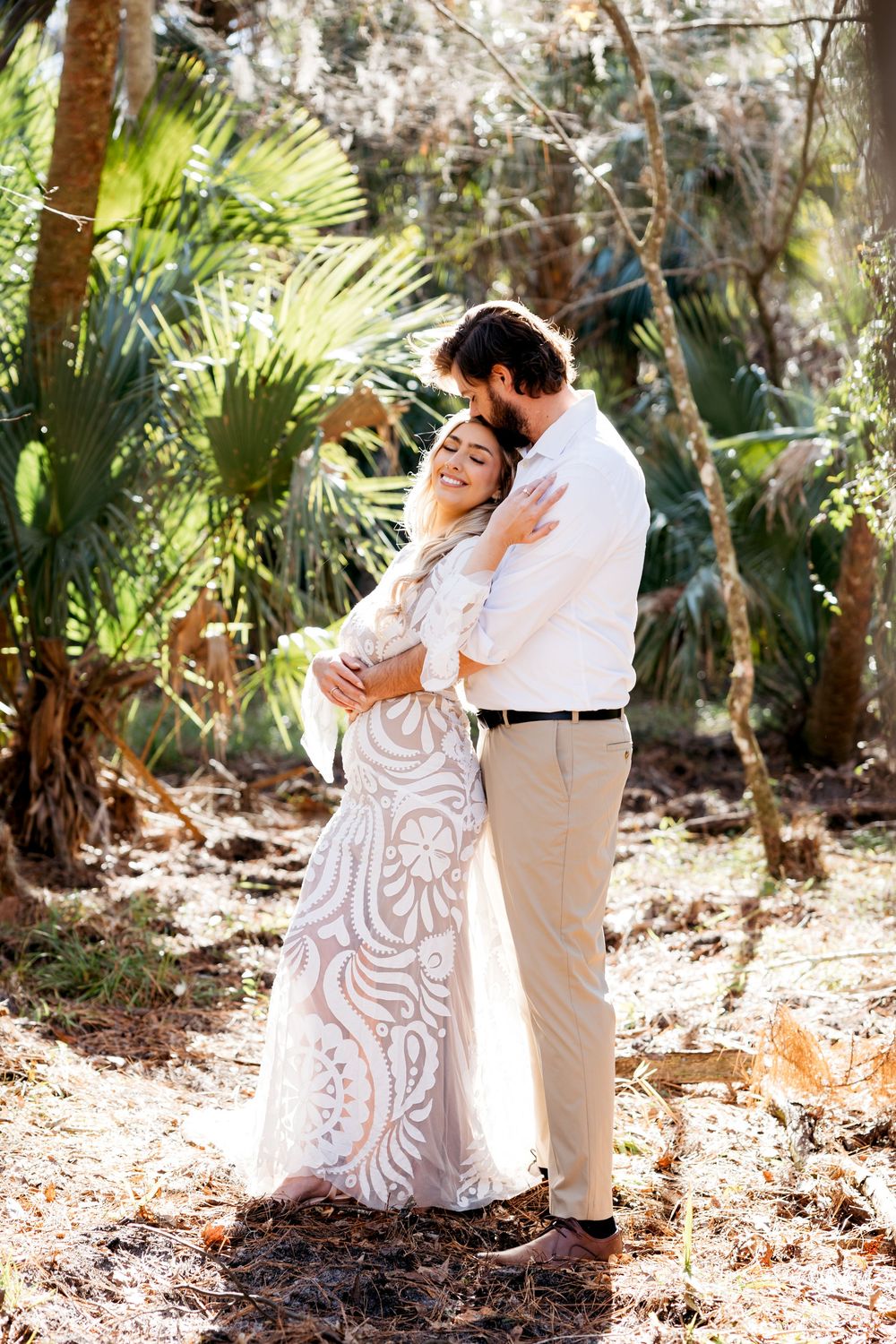 A loving couple embraces in a lush tropical garden setting, bride wearing an elegant white lace gown.
