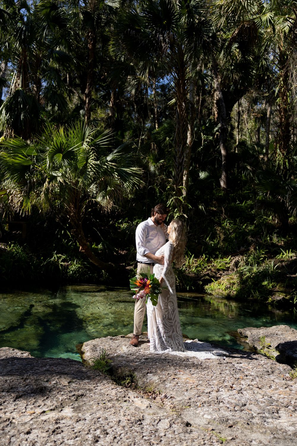 Two people embrace at a romantic natural springs setting surrounded by lush palm trees and tropical vegetation.
