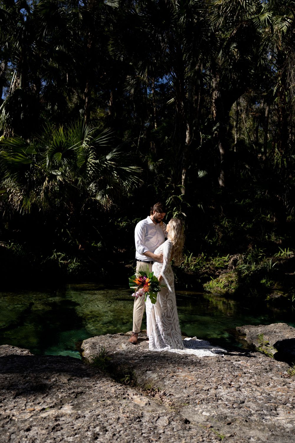 Couple stands together on a rock formation beside crystal clear spring waters surrounded by tropical palm trees and foliage.