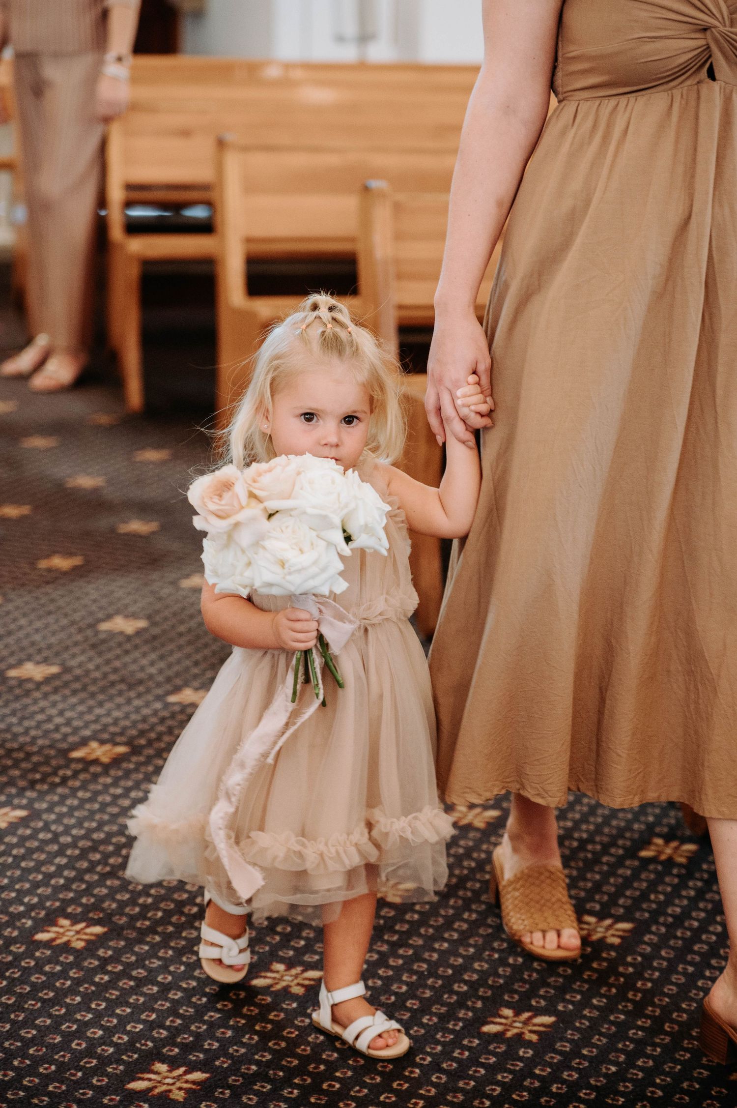 A young child in a beige tulle dress holds white roses while standing next to someone in a matching brown gown.