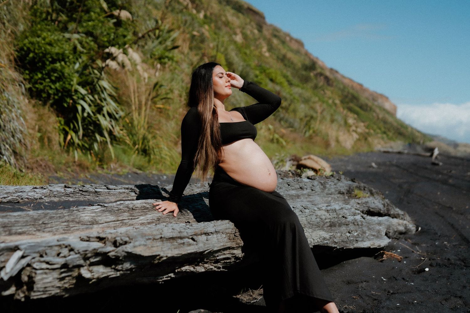 A maternity photo shoot on a rocky beach shows a person in a black flowing dress against a coastal backdrop.