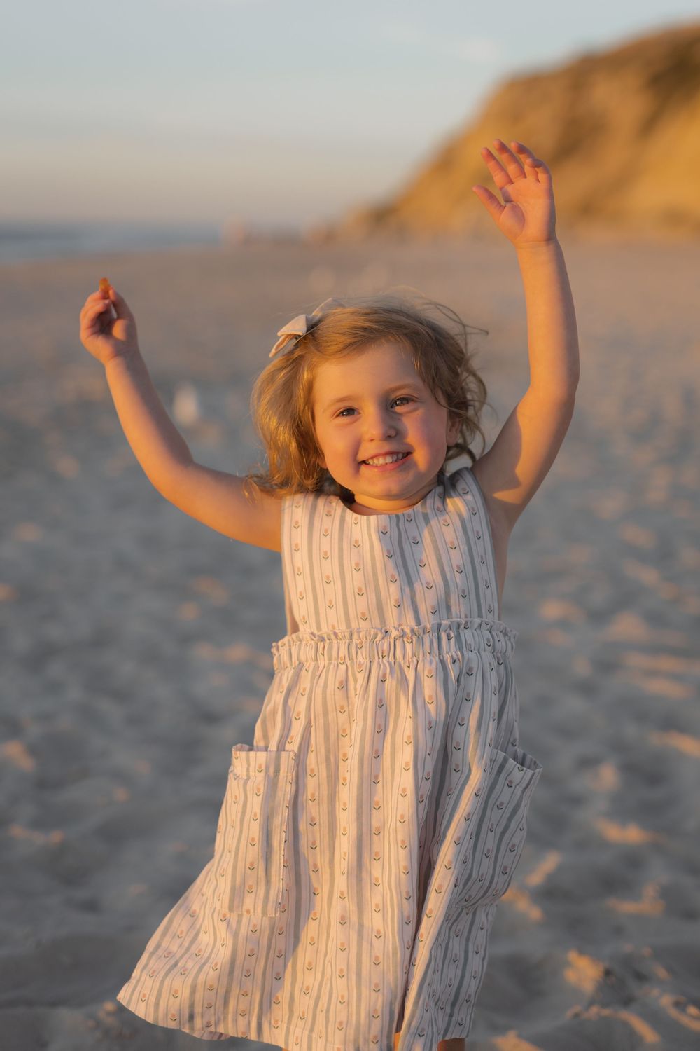 A small child in a white striped sundress plays joyfully on a sandy beach at sunset with cliffs in the background.