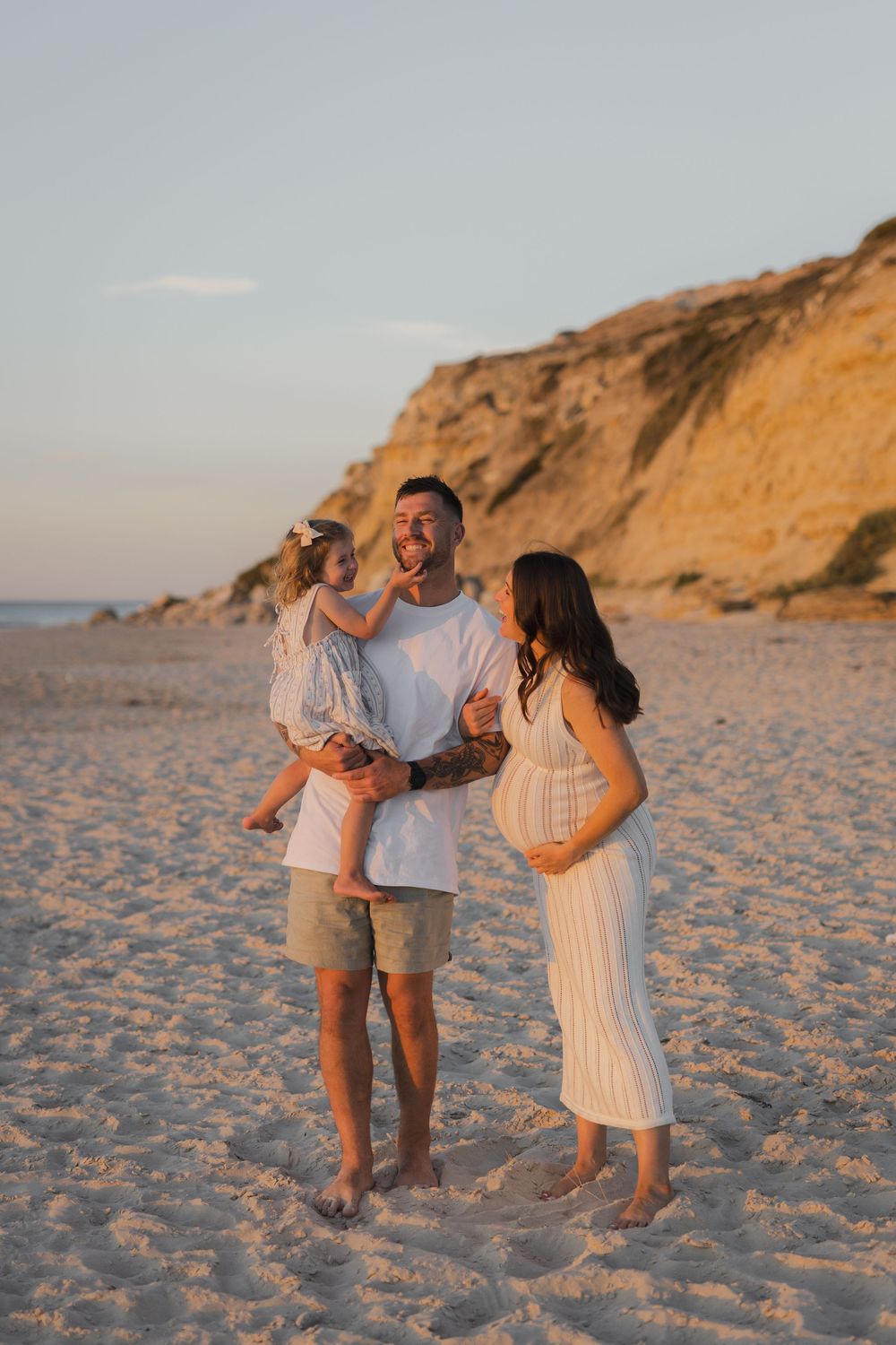 A family enjoys a sunset moment together on a sandy beach with towering golden cliffs in the background.