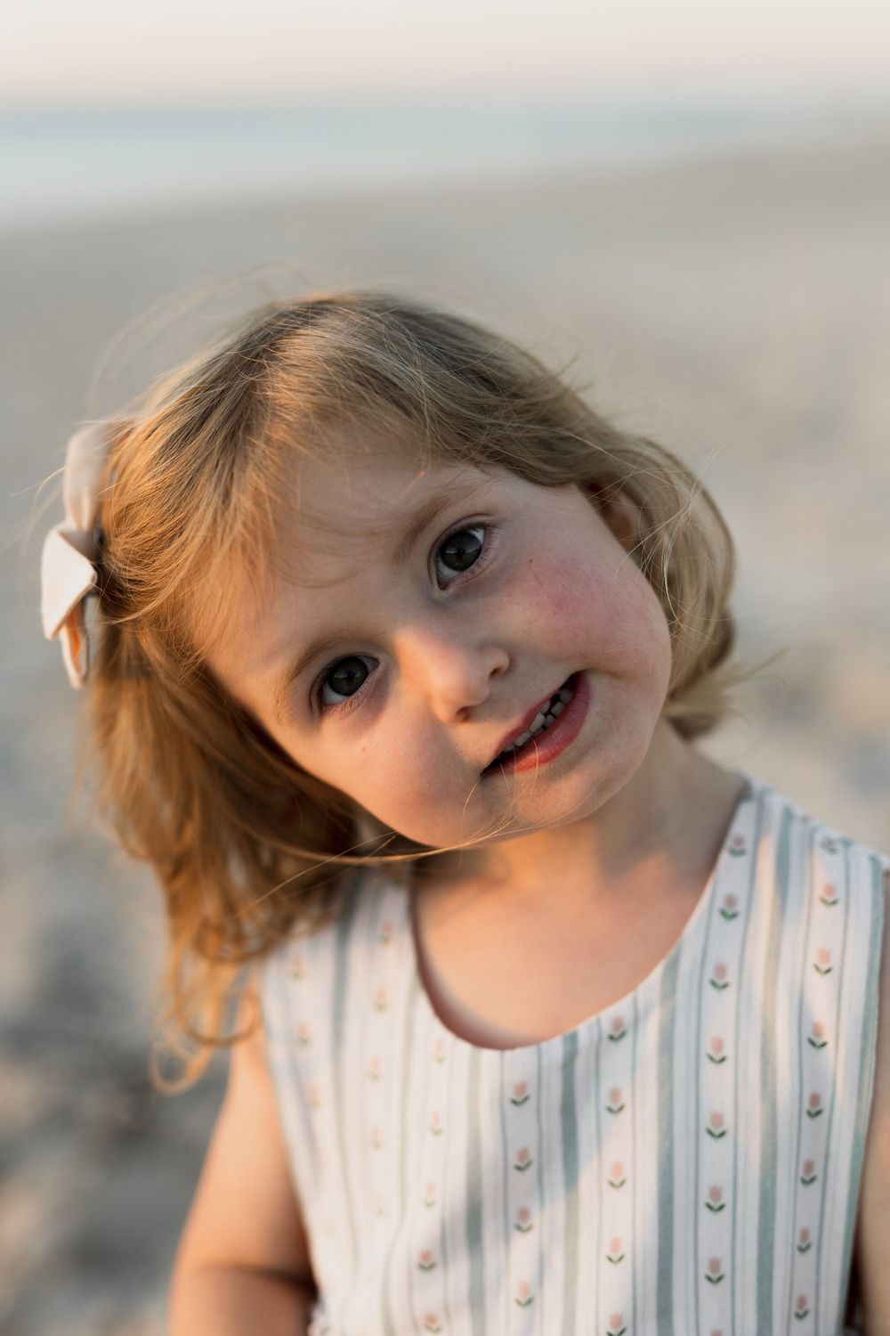 A young child with blonde hair in a white polka dot dress tilts their head and smiles at the camera in soft lighting.