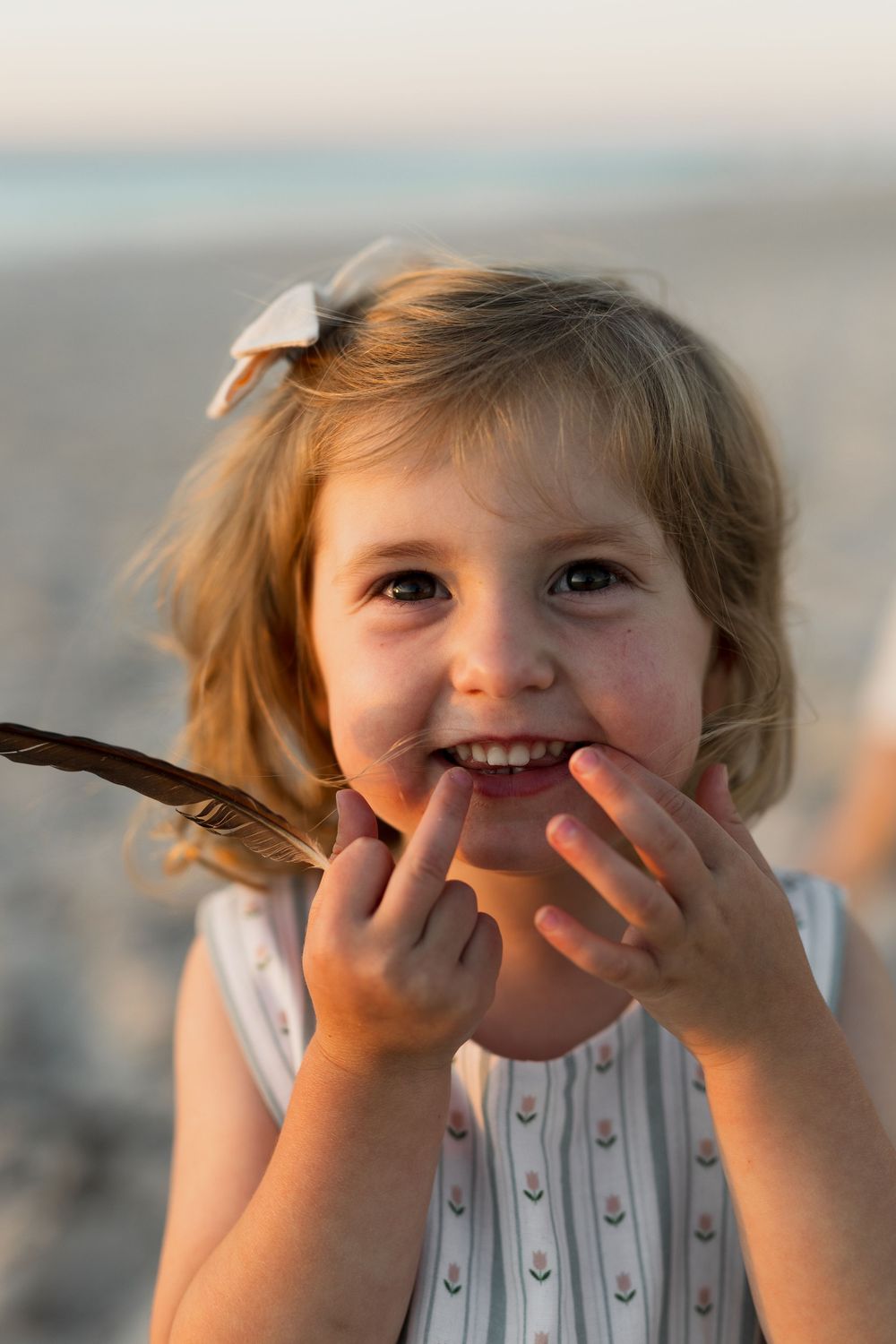 A child wearing a white dress smiles playfully while holding a brown feather at sunset.