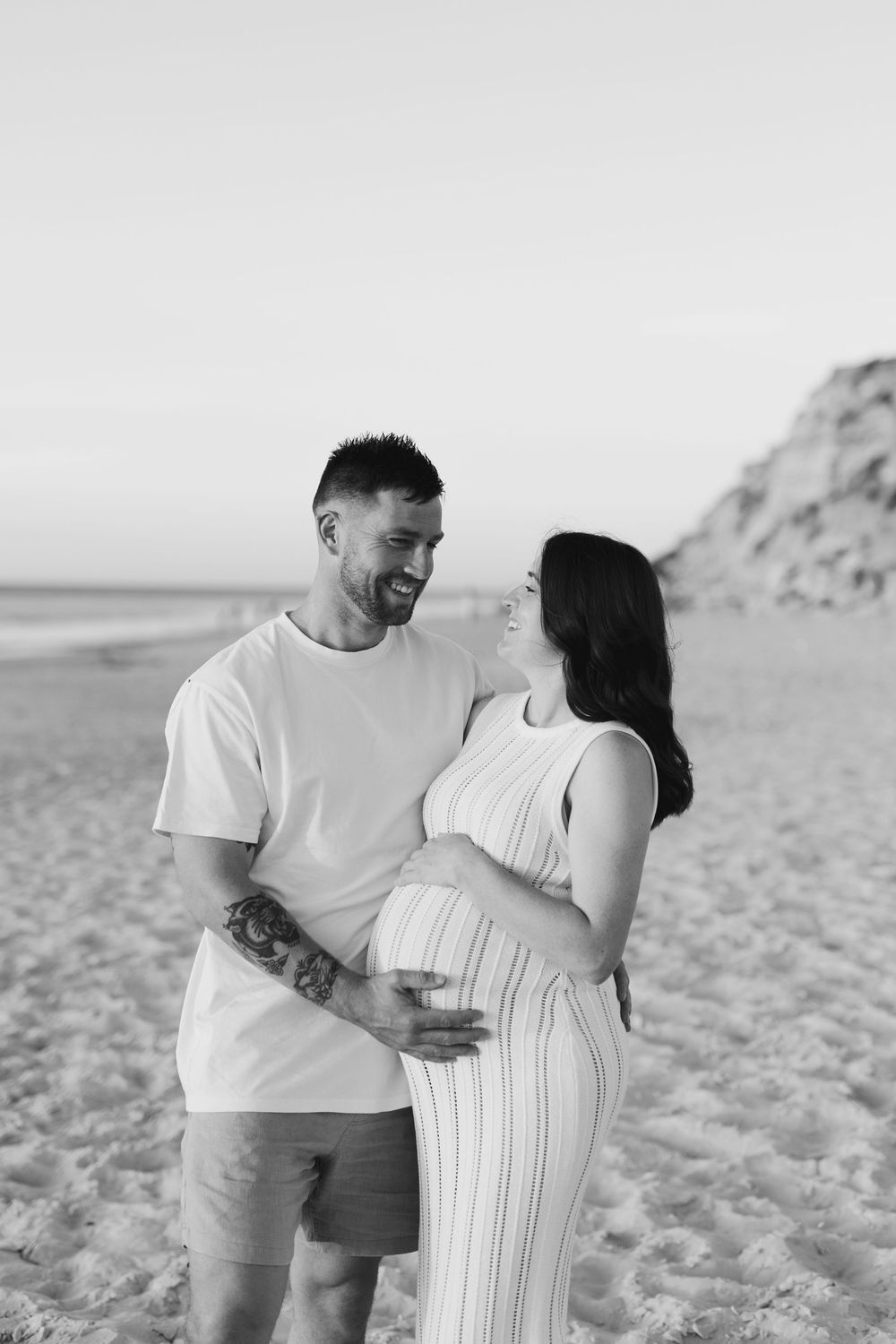 A couple shares an intimate moment on the beach as they celebrate their upcoming baby, the mother wearing a striped dress.