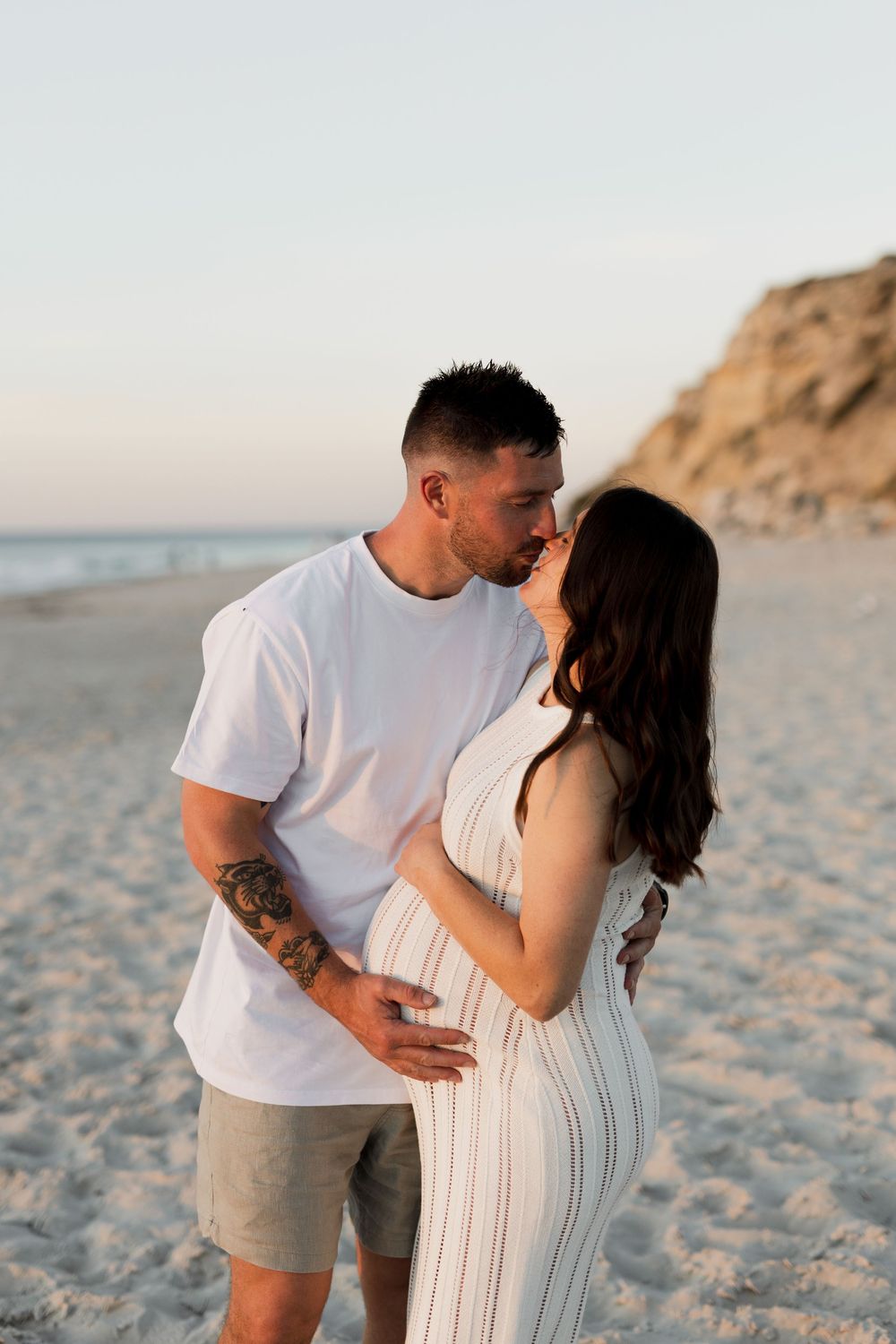 A couple embraces on a sandy beach at sunset with cliffs in the background.