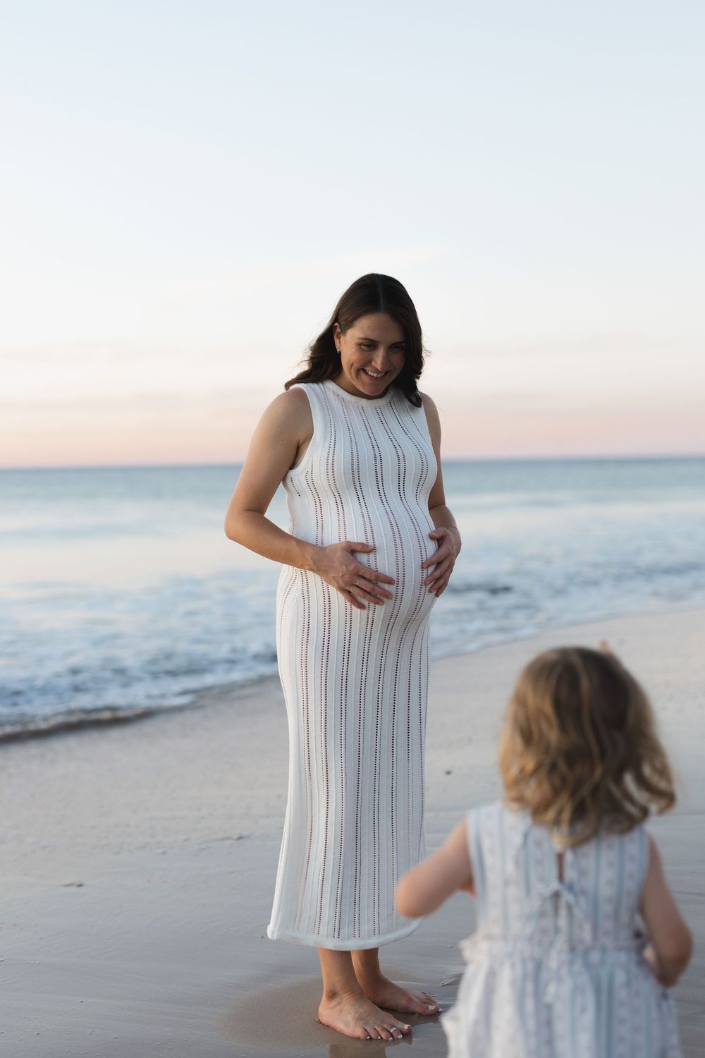 An expectant mother in a white ribbed dress shares a moment with a small child on a peaceful beach at sunset.