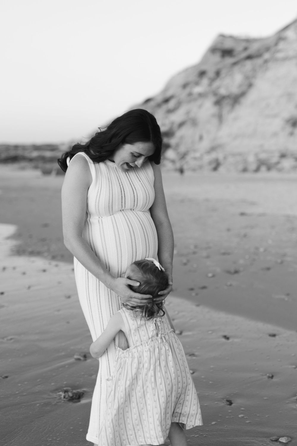 A pregnant woman in a white dress embraces a small child on a sandy beach with cliffs in the background.