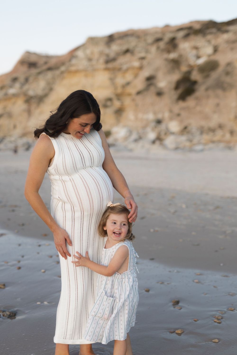 A heartwarming moment on the beach as mother in a white dress embraces her young daughter against a rocky cliff backdrop.
