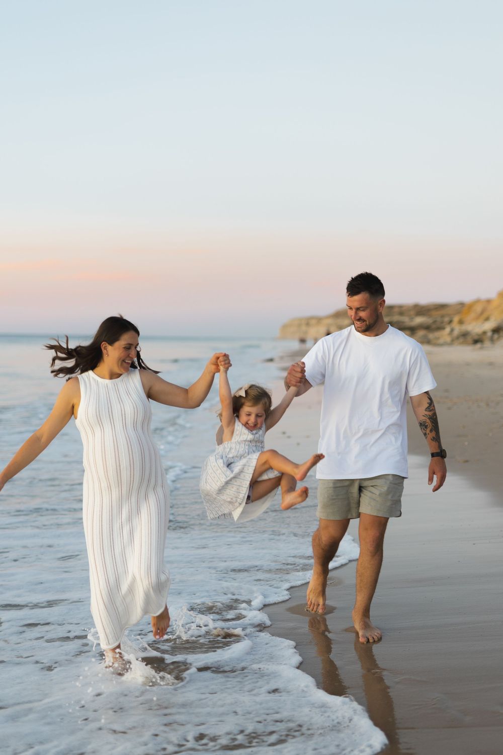 A family in white outfits swings their small child between them while walking barefoot along a sunset beach.