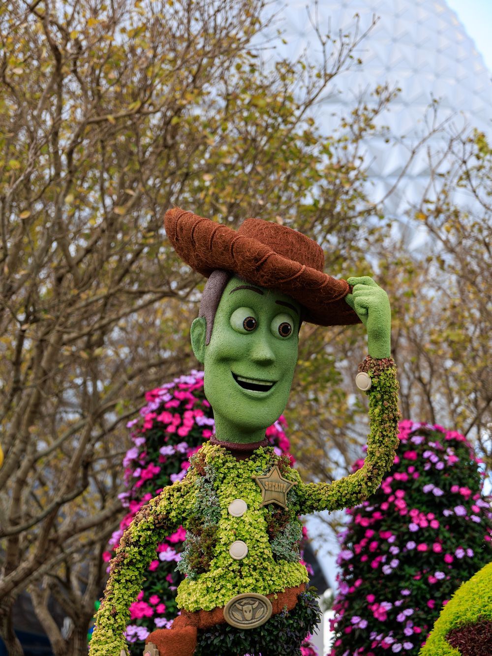 A Woody-themed topiary sculpture with brown hat and green moss body stands in front of pink flowers at a garden display.