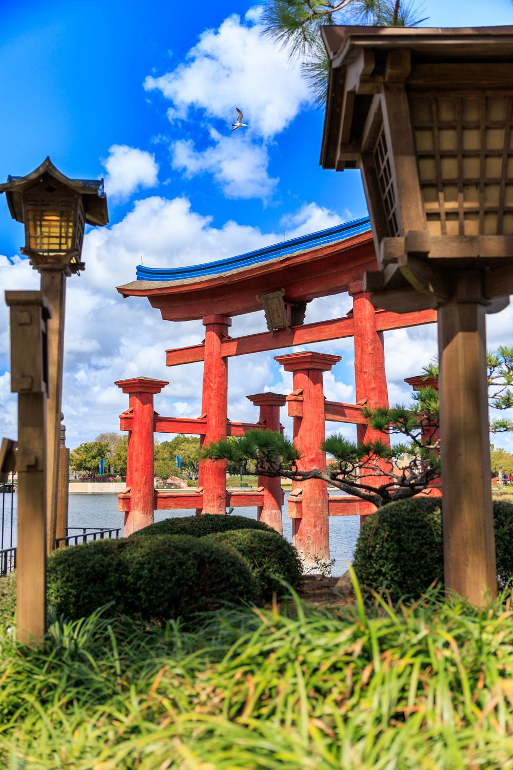 A row of traditional Japanese torii gates and lanterns against a blue sky with white clouds at a Shinto shrine.