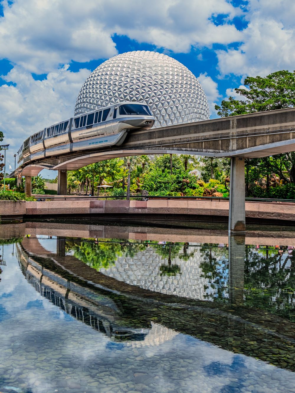 Disney World monorail track reflects in water with Spaceship Earth dome and blue sky visible.