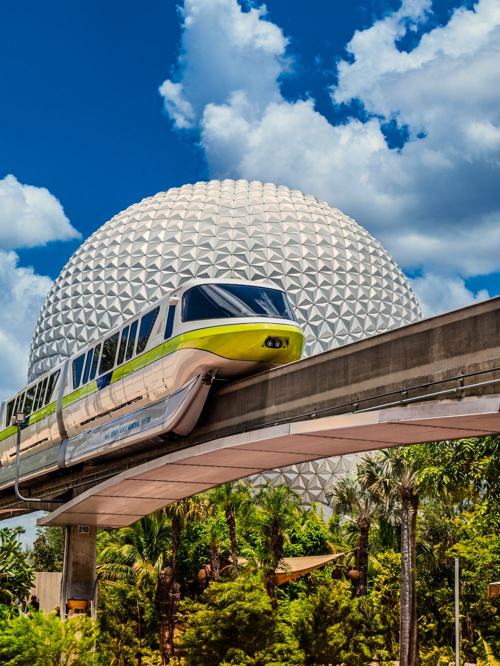 Yellow and white monorail glides past Spaceship Earth sphere at EPCOT under blue skies.