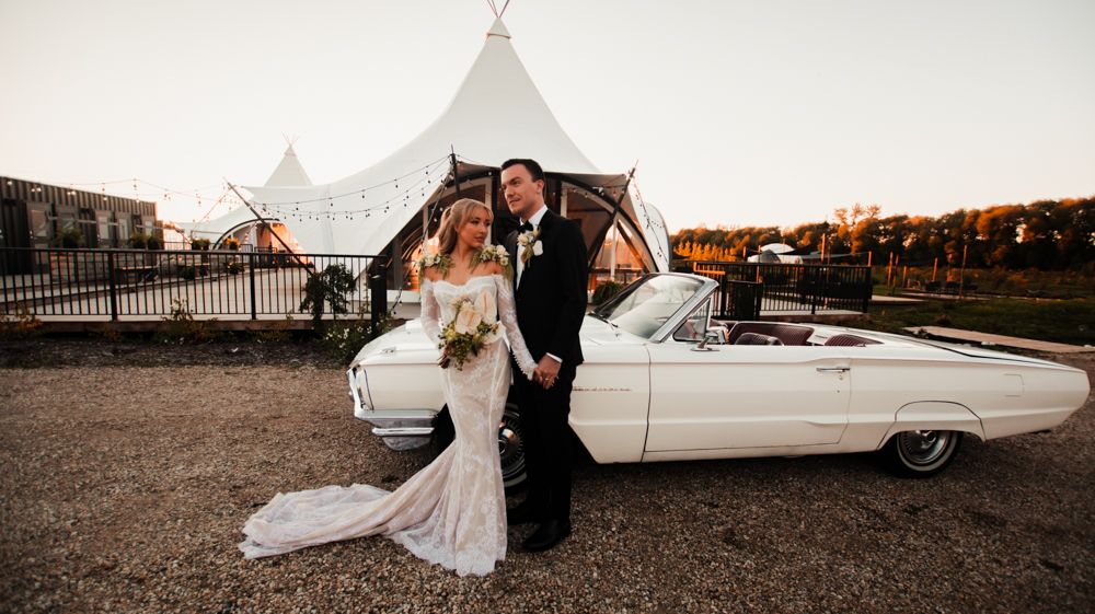 Newlyweds pose beside classic white Cadillac convertible at sunset with white tent pavilions in background.