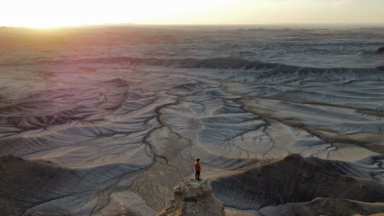 A silhouetted figure stands on a cliff edge overlooking dramatic desert canyons at sunset.