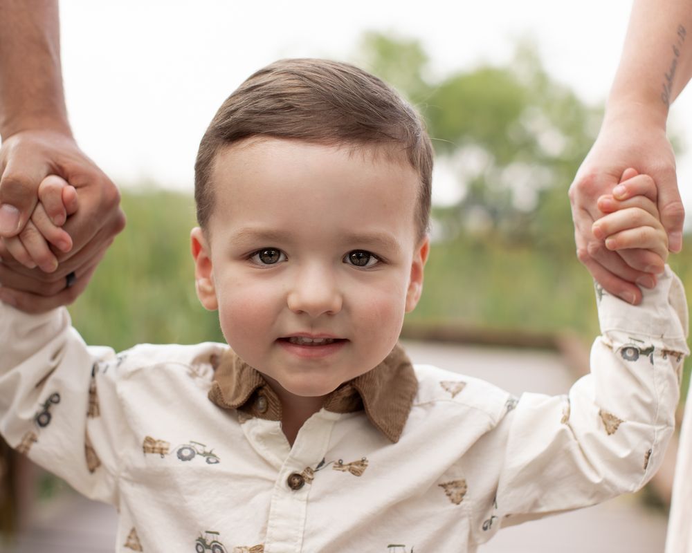 A toddler in a beige button-up shirt smiles while being held by adult hands against a blurred outdoor background.