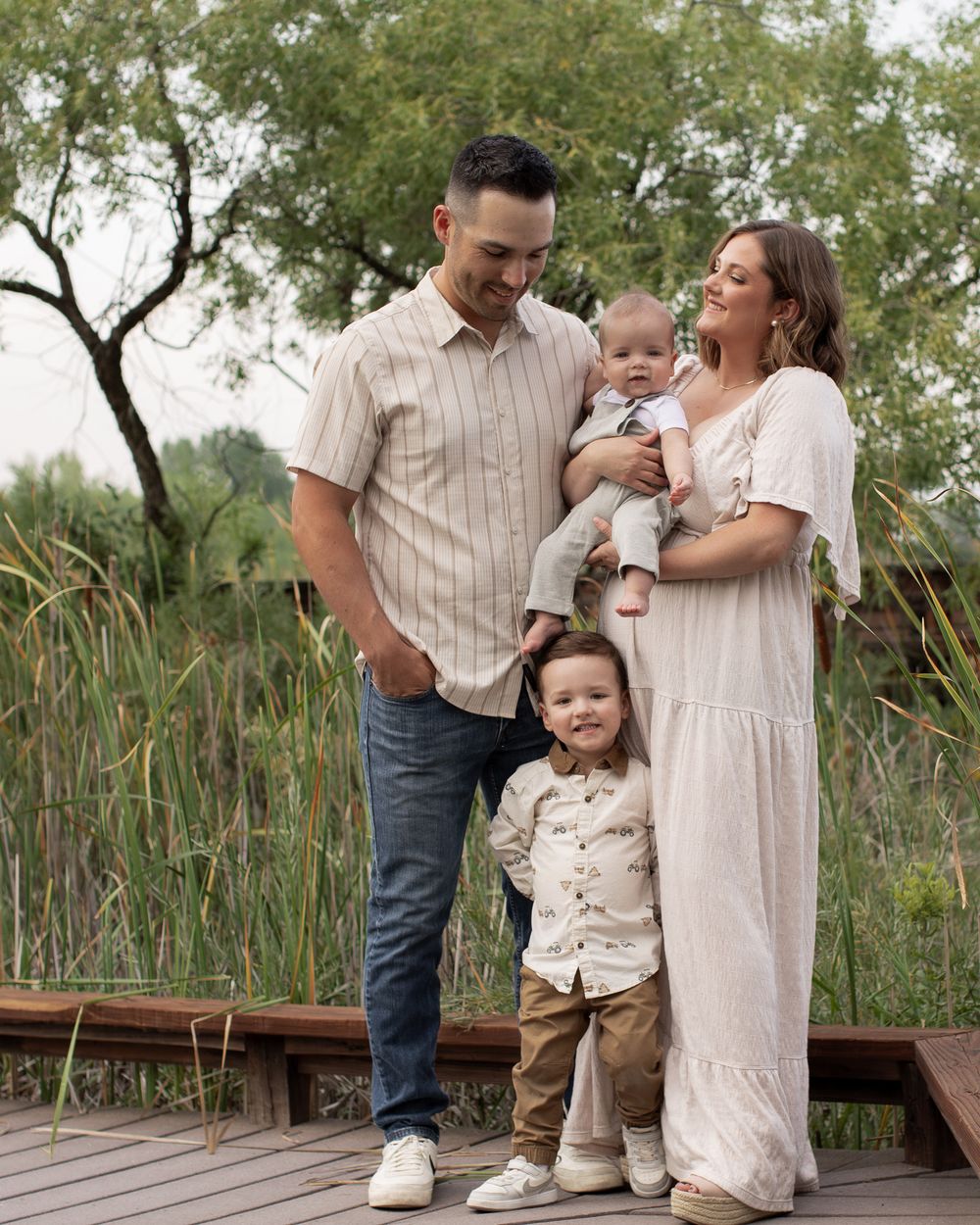 Family shares tender moments together on wooden boardwalk surrounded by tall grass and trees.