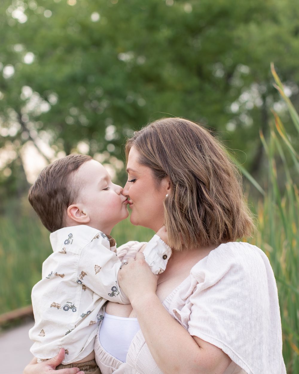 Mother and child share sweet kisses outdoors in natural setting with bokeh background.
