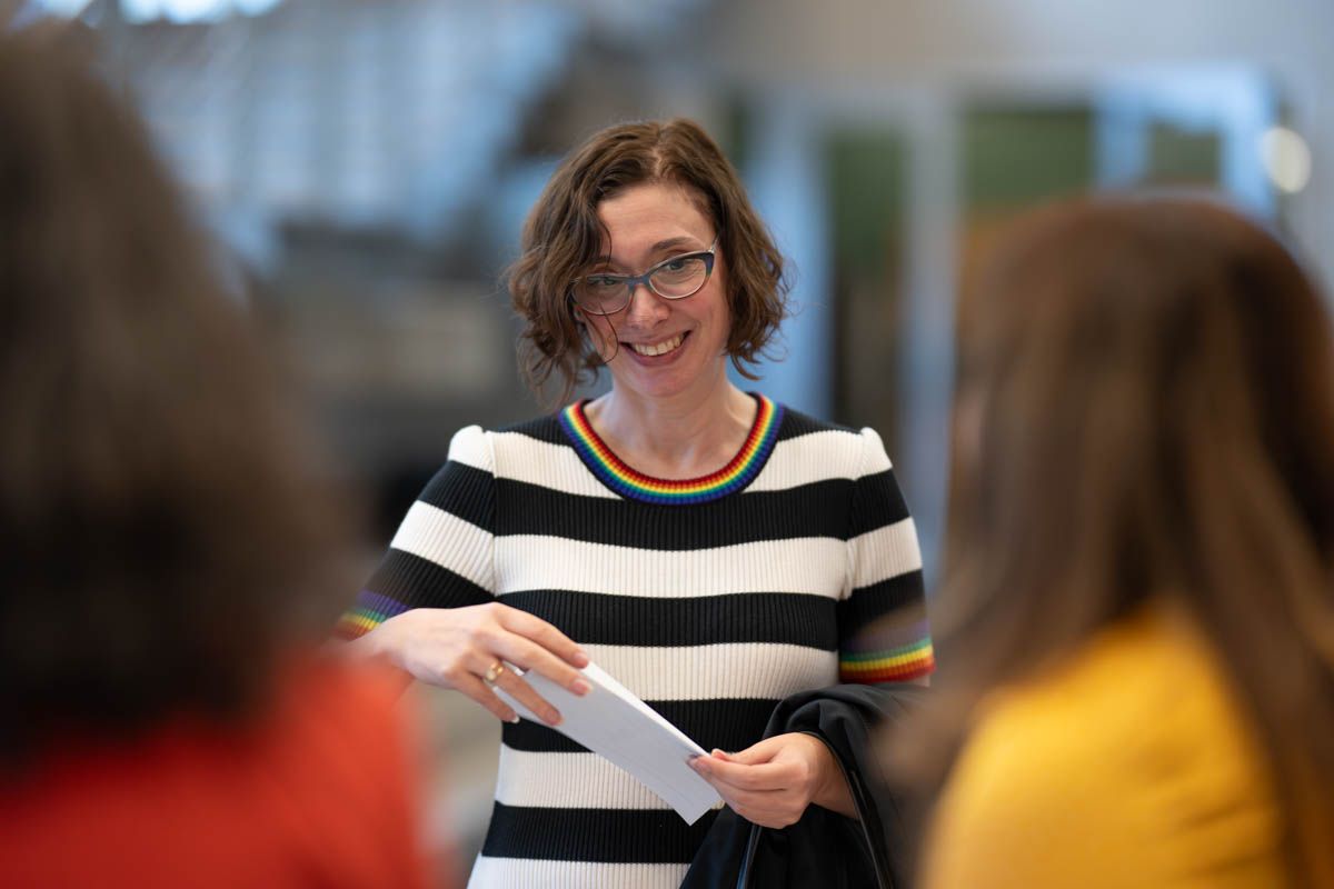 A person in a black and white striped shirt smiles while holding papers in an office setting.