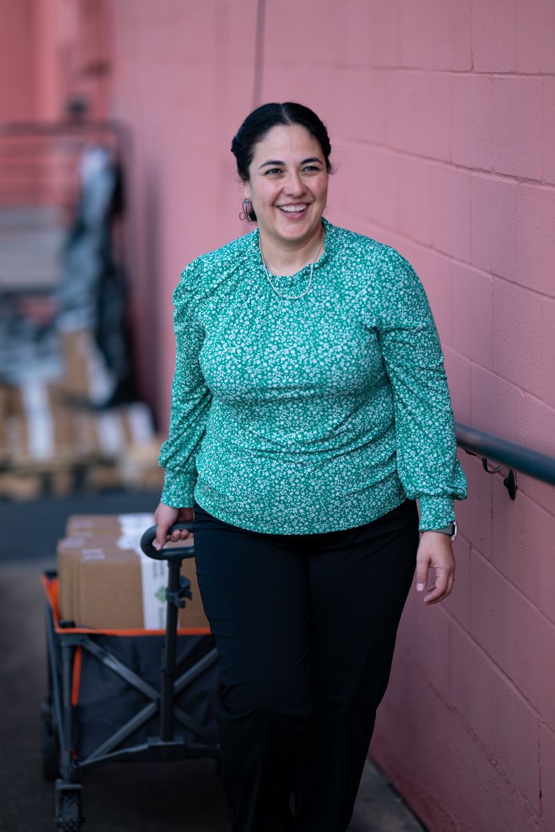 A person wearing a teal polka dot blouse and black pants poses against a pink wall in a casual setting.