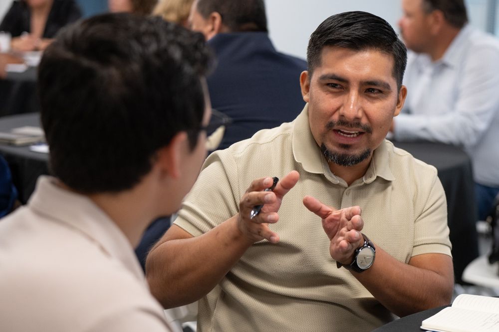 A person in a beige shirt smiles and gestures while engaged in conversation at a table.